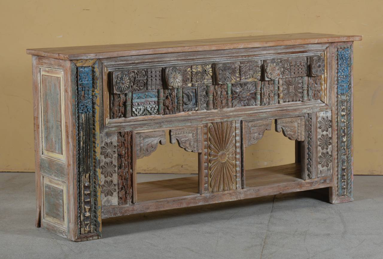 An ornate carved wooden sideboard or console table with distressed blue, beige, and brown painted details, set against a plain beige wall and gray floor.