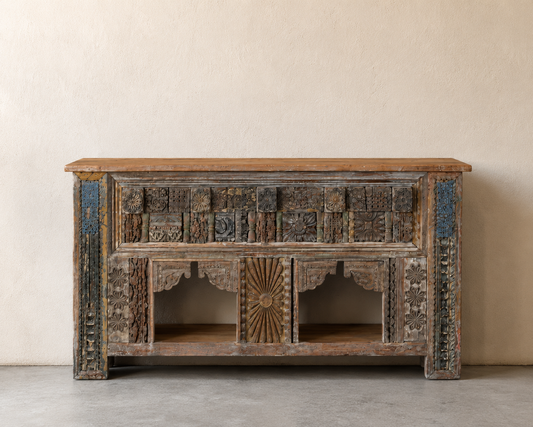 An ornate carved wooden sideboard or credenza with a warm brown wood top, distressed dark wood body, and blue painted accents on the side panels, set against a neutral beige wall and gray floor.