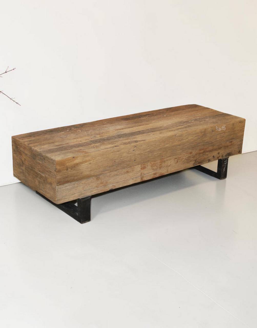 A rustic rectangular wooden bench with a thick natural wood top and black metal support legs, photographed against a plain white wall and floor.