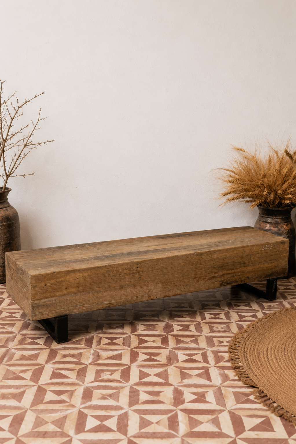 A rustic rectangular wooden bench with black metal legs, placed on a patterned terracotta tile floor against a plain white wall, styled with dried plants in rustic vases.