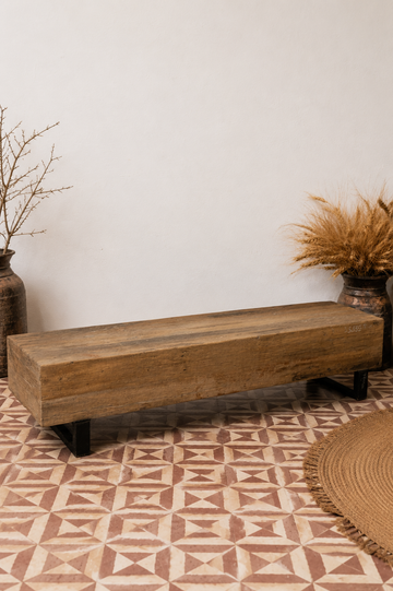 A rustic rectangular wooden bench with black metal legs, placed on a patterned terracotta tile floor against a plain white wall, styled with dried plants in rustic vases.