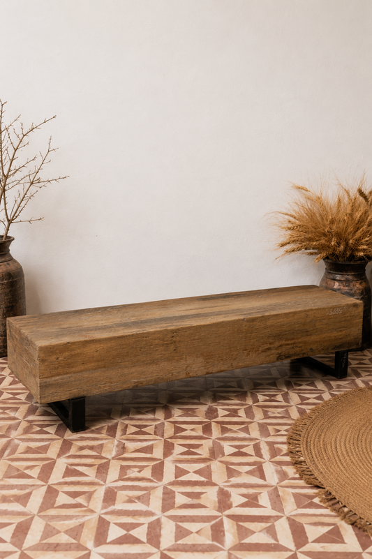 A rustic rectangular wooden bench with black metal legs, placed on a patterned terracotta tile floor against a plain white wall, styled with dried plants in rustic vases.