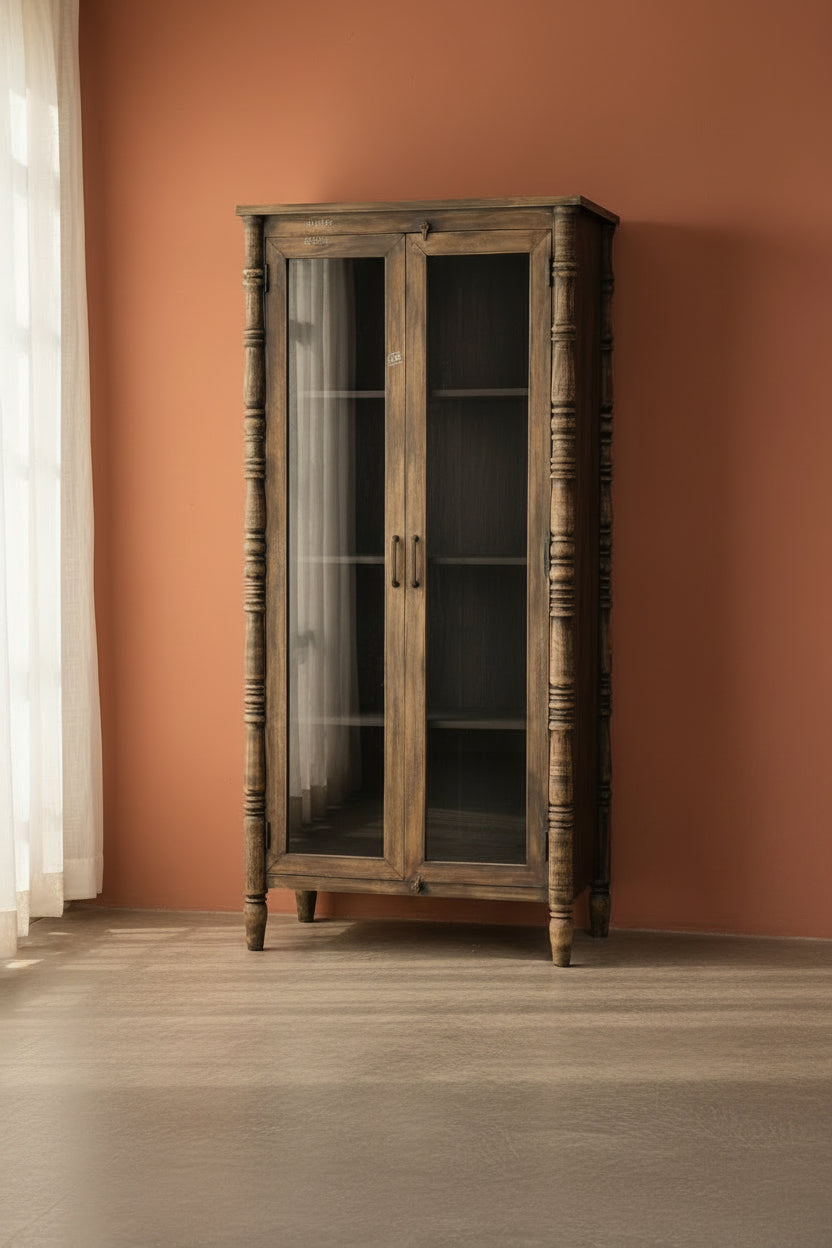 A rustic dark brown wooden bookcase cabinet with glass doors, turned spindle legs, and open shelves, set against a plain light gray wall and concrete floor.