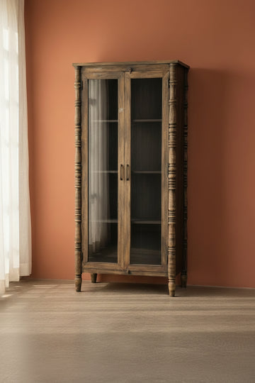 A rustic dark brown wooden bookcase cabinet with glass doors, turned spindle legs, and open shelves, set against a plain light gray wall and concrete floor.