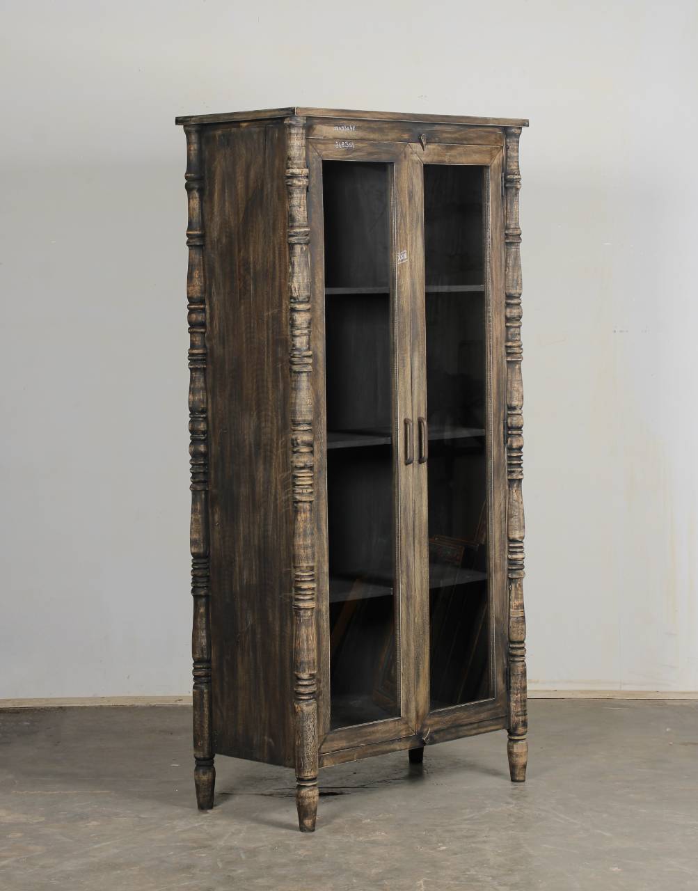 A rustic dark brown wooden bookcase cabinet with glass doors, turned spindle legs, and open shelves, set against a plain light gray wall and concrete floor.