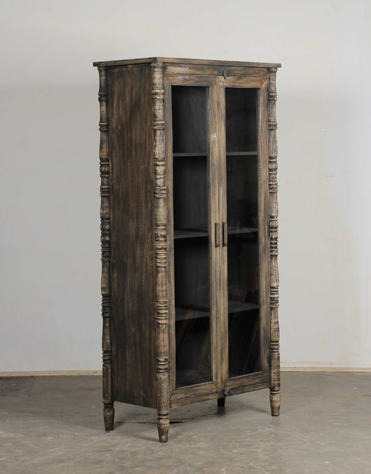 A rustic dark brown wooden bookcase cabinet with glass doors, turned spindle legs, and open shelves, set against a plain light gray wall and concrete floor.