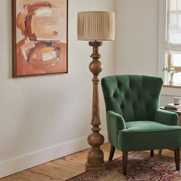 Wooden floor lamp with pleated shade next to a gray armchair in a room with a painting on the wall.
