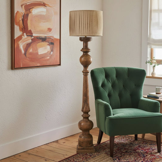 Wooden floor lamp with pleated shade next to a gray armchair in a room with a painting on the wall.