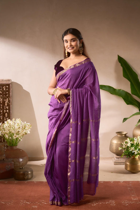 Woman in a purple saree standing indoors with decorative elements.