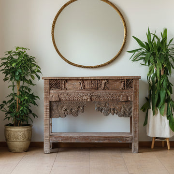 Decorative wooden console table with intricate carvings, round mirror, and potted plants in a room.