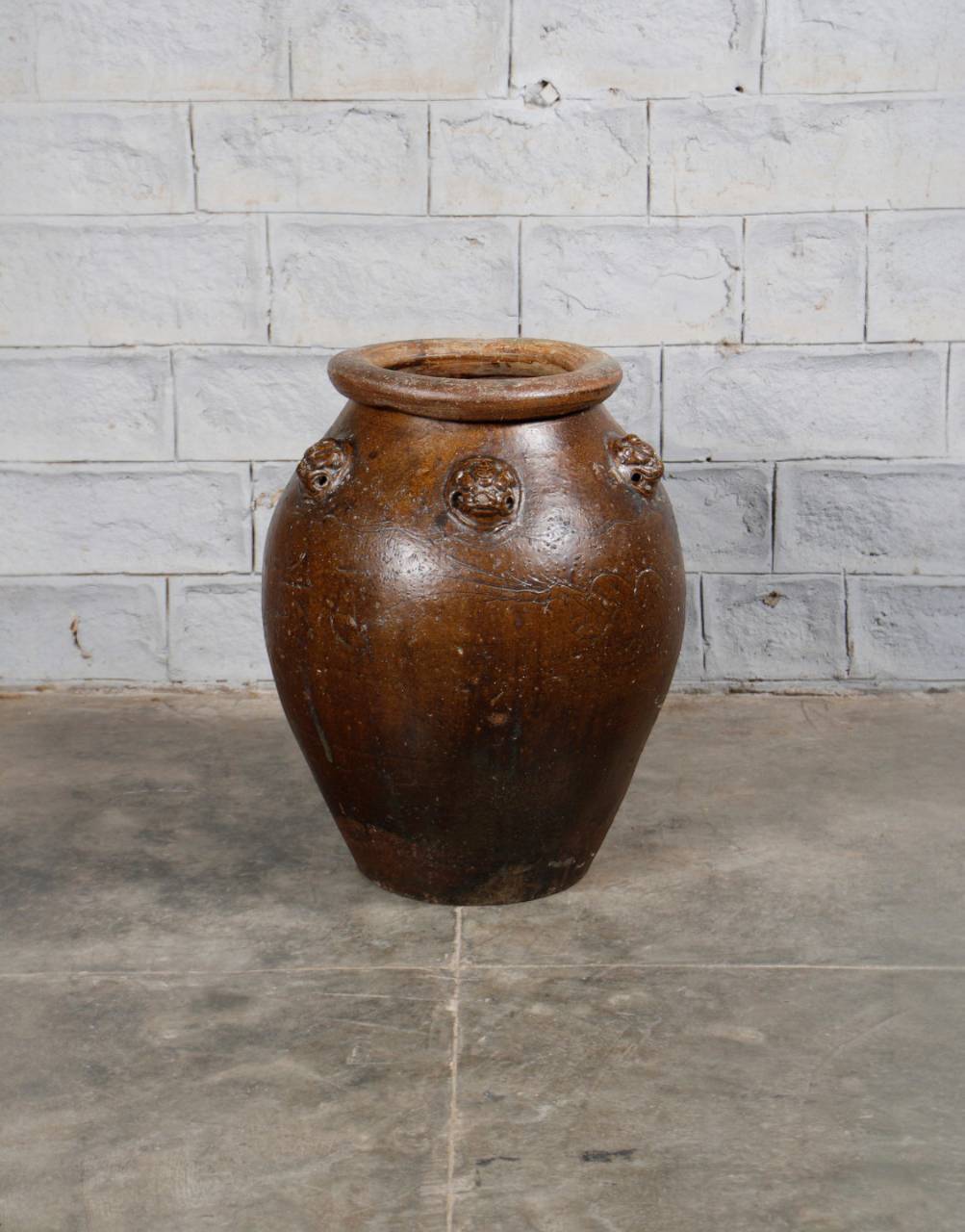 Brown ceramic jar with lion head designs on a stone floor against a stone wall.