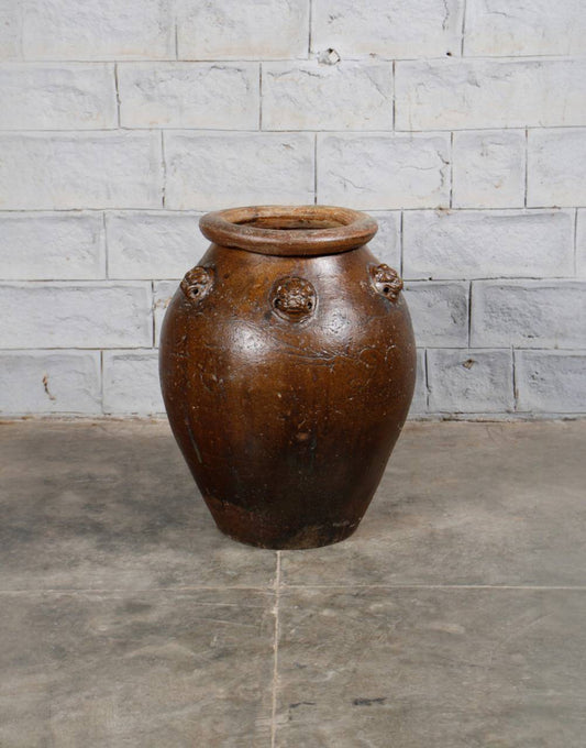 Brown ceramic jar with lion head designs on a stone floor against a stone wall.