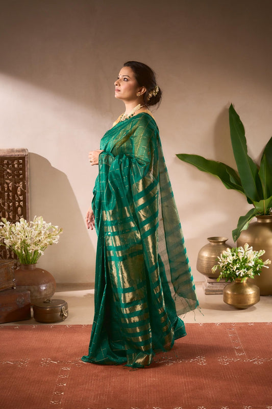 Woman in a green saree standing in a room with decorative vases and plants.