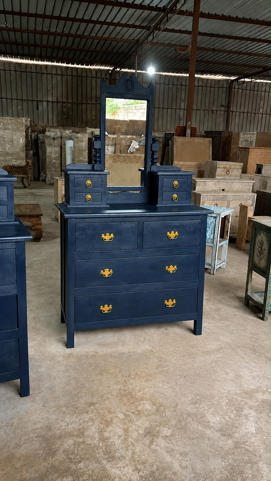 A dark blue wooden dresser with a matching mirror and side tables, featuring gold-colored ornate handles, photographed in a warehouse setting.