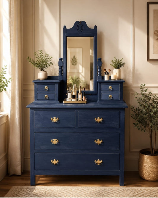 A dark blue wooden vanity dresser with a matching mirror, ornate gold handles, and decorative side tables, styled in a warm beige room with plants.