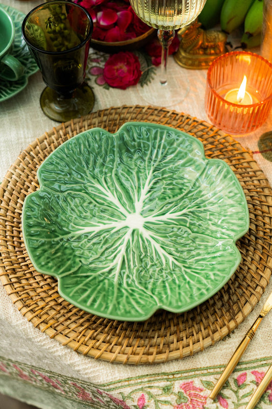 Green leaf-shaped ceramic plate on a woven placemat with candles and flowers in the background.