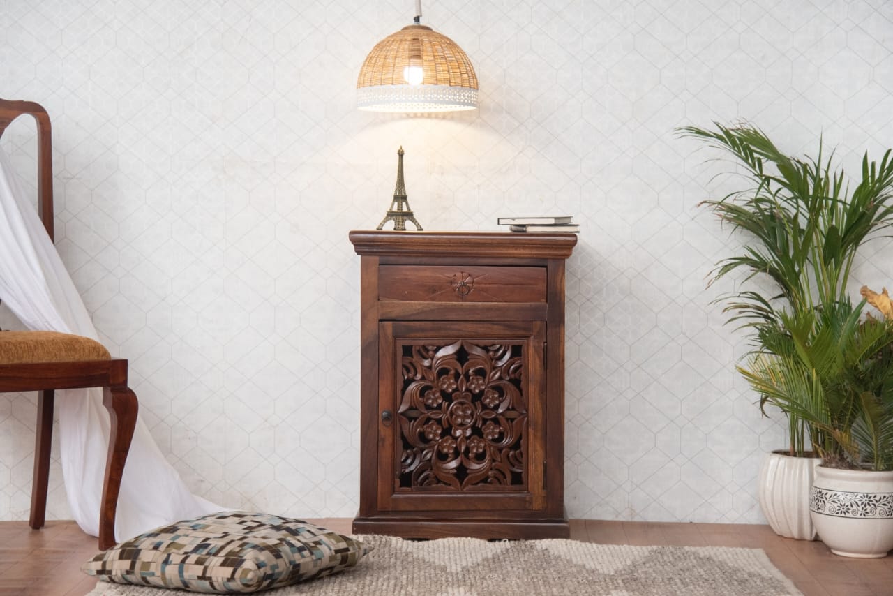 Wooden side table with intricate carvings in a room with a chair, lamp, and plant.