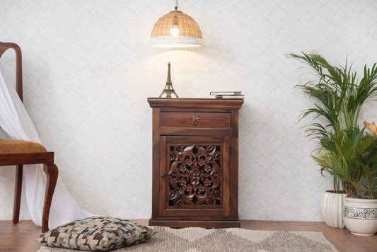 Wooden side table with intricate carvings in a room with a chair, lamp, and plant.