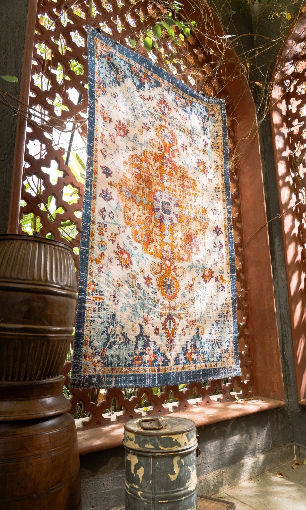 Decorative rug with intricate patterns on a wooden stand, surrounded by plants and a barrel.