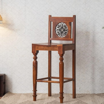 Wooden bar stool with decorative backrest in a room setting with a lamp and plant.