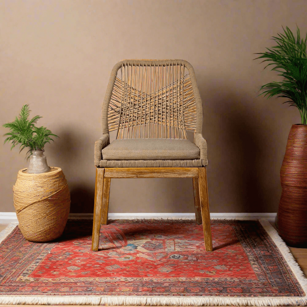 Wicker chair with a cushion on a red patterned rug against a beige wall, with two plant pots on either side.