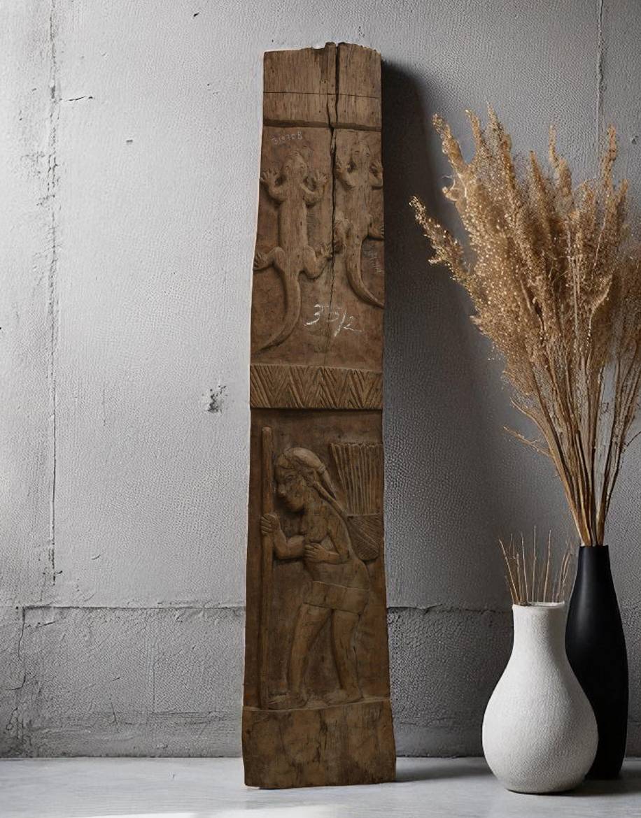 Wooden panel with carvings next to a vase with dried plants against a gray wall.
