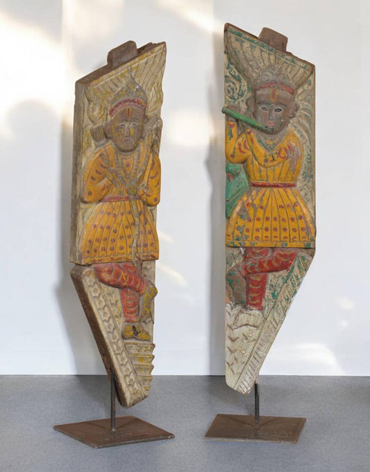 Two wooden carvings of figures on stands against a white wall.
