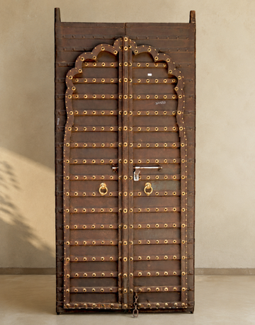 A tall, narrow dark brown wooden door with an arched top and heavily decorated with brass-colored metal studs, rings, and a central handle, set against a neutral beige wall.