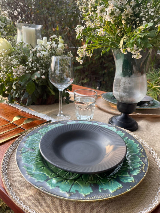 Dining table setting with dark gray plates, green leafy placemats, and clear glasses on a natural outdoor background.