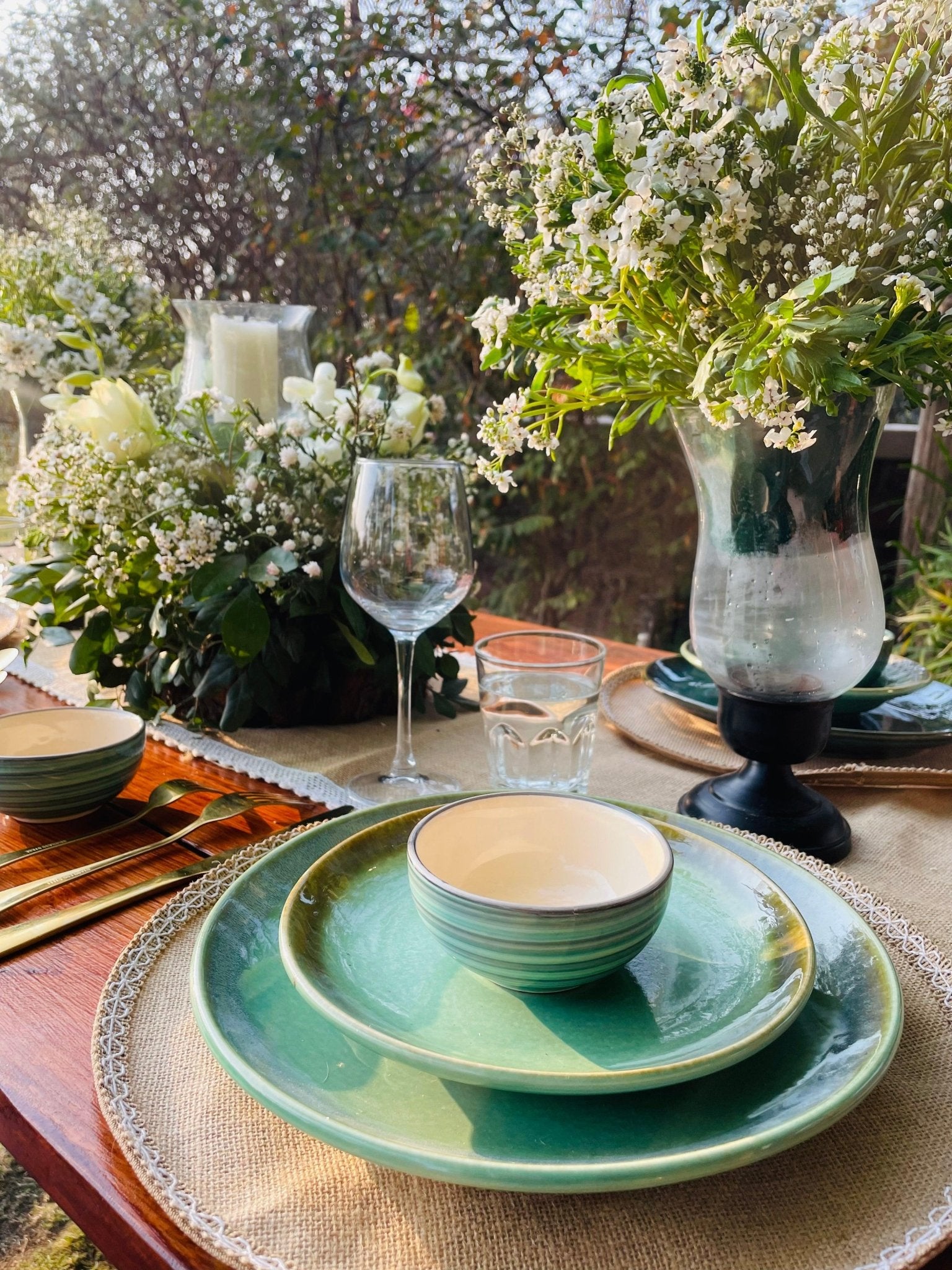 Table setting with green ceramic plates and bowls, glasses, and floral arrangements outdoors.