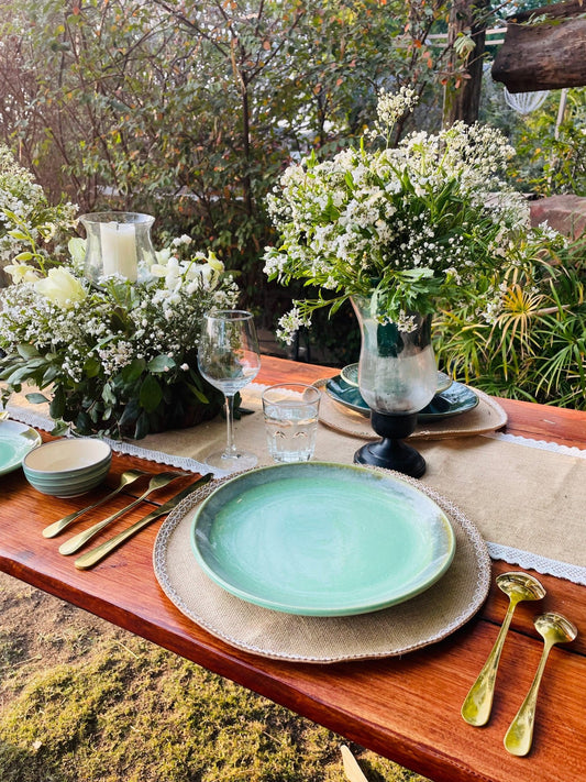 Dining table set with green plates, gold cutlery, and white flowers outdoors.