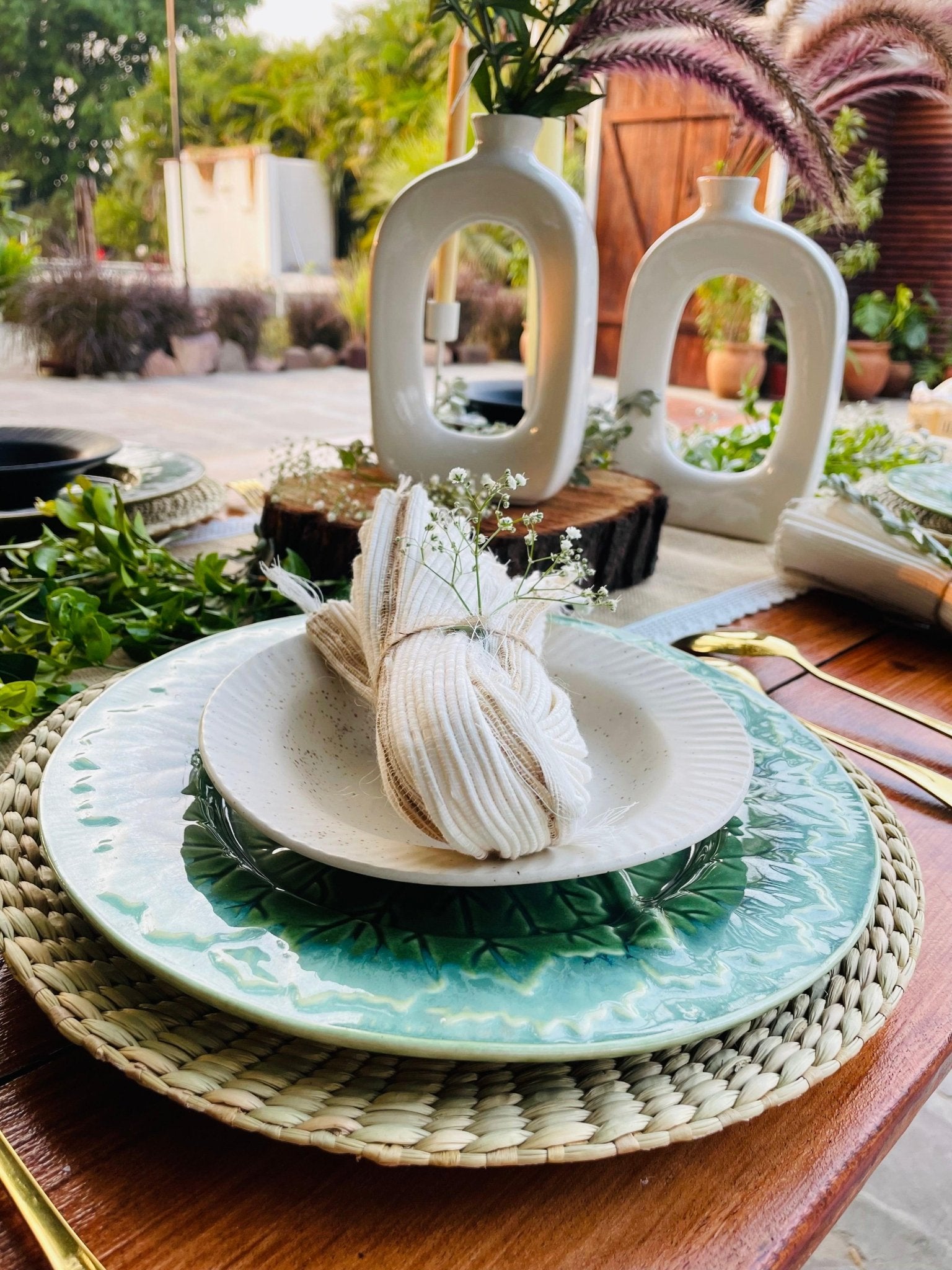 Dining table setting with a folded napkin on a plate, surrounded by greenery.