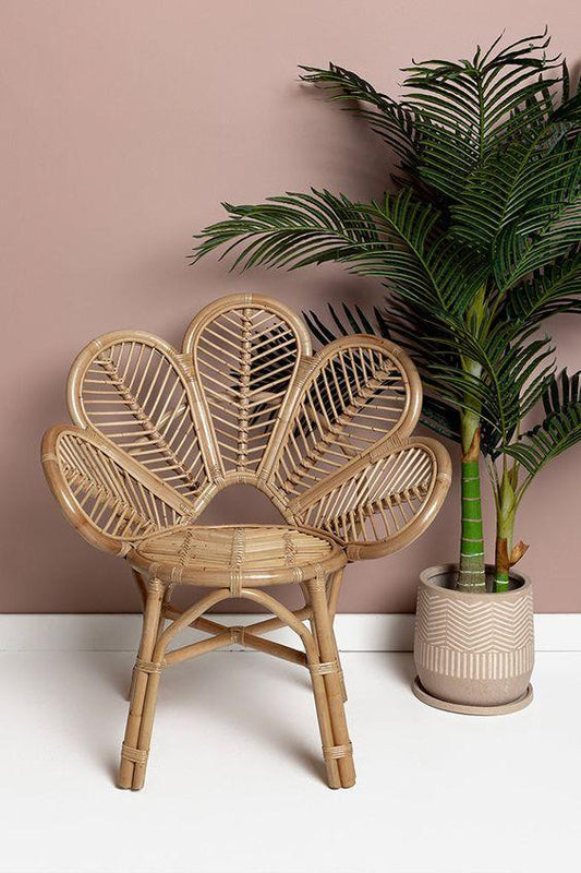 Wicker chair with leaf-patterned backrest next to a potted plant on a white surface with a pinkish-beige wall background.